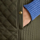 Close-up of a hand in a quilted olive-green gilet pocket, with a branded button and a blue ribbed sleeve visible.