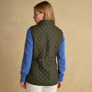 A person wearing a green quilted gilet over a blue jumper, with beige striped shorts, poses against a textured beige background.