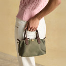 A woman in a pink sweatshirt holds a small khaki green handbag with brown leather accents, against a soft beige backdrop.