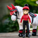 Toy doll in a red riding outfit and helmet stands beside a white toy horse with red gear. Greenery is blurred in the background. Equestrian theme.