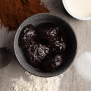 A bowl of dark chocolate pudding on a grey surface, surrounded by piles of cocoa powder, flour, sugar, and a dish of cream, conveying a rich, indulgent tone.