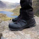 Black and dark grey mid-height hiking boots with red detailing and rugged grip soles, worn on rocky mountain terrain overlooking a lake.