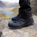 Black and dark grey mid-height hiking boots with red detailing and rugged grip soles, worn on rocky mountain terrain overlooking a lake.