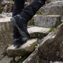 Rear three-quarter view of black hiking boots stepping up rugged stone terrain on a mountain trail.