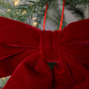 A large, plush red velvet bow hangs on a Christmas tree. The bow is vibrant against the backdrop of green pine needles and soft glowing lights.