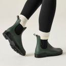 Rear three-quarter view of dark green ankle wellington boots worn with white socks and black leggings, highlighting the rugged tread soles.