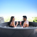 Smiling couple relaxing in the MSpa Mono-Eco inflatable hot tub, enjoying drinks while surrounded by bubbling water and a scenic outdoor view.