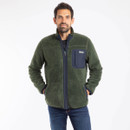 A man stands confidently wearing a green fleece jacket with a navy pocket over a black t-shirt, paired with jeans, against a plain white background.