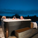 A smiling couple toasting with champagne glasses in the MSpa Oslo Plus 6 Person Hot Tub at night, set on a wooden deck beside a tranquil lake.