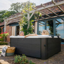 A couple relaxing in the MSpa Oslo Plus Hot Tub, 6 Person, set on a stone patio in a garden with blooming roses and a wooden pergola in the background.
