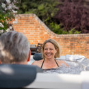 A smiling woman relaxing in the MSpa Oslo Plus 6 Person Hot Tub, chatting with a companion, surrounded by bubbling water and a peaceful garden setting.