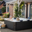 A couple relaxing in the MSpa Oslo WiFi Controlled 6 Person Hot Tub in a sunny garden, surrounded by blooming flowers and a rustic brick house.