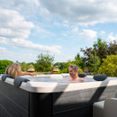 A couple smiling and chatting while enjoying the MSpa Oslo Plus 6 Person Hot Tub in a lush garden, surrounded by greenery under a bright blue sky.