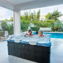 A smiling couple enjoying the MSpa Oslo Sapphire 4–6 Person Hot Tub indoors, toasting with drinks while surrounded by bubbling water and a view of a pool and garden.