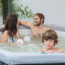 Close-up of a family relaxing in an MSpa hot tub — a man and woman smile at each other while a young boy leans on the spa edge, with soft glowing lights floating in the bubbling water.