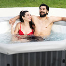 A smiling couple relax together in a bubbling MSpa hot tub set in a garden, enjoying a warm soak with arms resting on the cushioned edge.