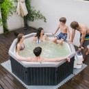 A group of five people, including two adults and three children, enjoy a relaxing soak in an octagonal MSpa hot tub on a garden deck, while one adult adjusts the spa settings using the control unit.