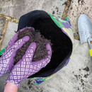Hands in purple gloves gather dark soil from a green and purple bag placed on stone tiles. A small silver trowel lies nearby, surrounded by scattered soil.