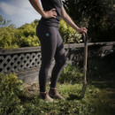 A person stands confidently beside a spade in a garden, wearing dark leggings and boots. Sunlit greenery and a lattice fence are in the background.