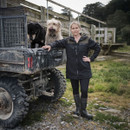 A person in a black jacket and boots stands confidently by a muddy off-road vehicle with two dogs on it, set against a rural backdrop with a wooden fence.
