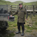 Man in a green jacket and black boots stands beside a quad bike on a farm, with grassy hills in the background. He appears focused and ready for work.