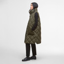 A person stands in profile, wearing a long, quilted olive green vest over a black outfit. The setting is minimal, conveying a modern, stylish tone.