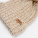 A beige, ribbed knit beanie with a brown leather tag displaying "Barbour" is shown on a light background. The texture looks soft and cosy.