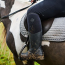 Muck Boots Women's Colt Ryder All-Conditions Riding Boot in use, shown muddy on a rider’s foot in stirrups while seated on a black and white horse.