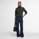 A person stands confidently wearing a dark green turtleneck sweater and wide-leg blue jeans. They hold a black handbag and have braided hair.