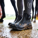 Close-up of Muck Boots Wear Ankle Wellington Boots in black, shown muddy and worn on a person standing on wet ground near horses, highlighting durable build and low-cut design.