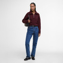 A person stands confidently against a plain background, wearing a burgundy sweater, blue jeans, and black shoes, with a black shoulder bag. The mood is casual.