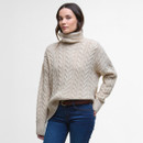 A woman in a cream cable-knit turtleneck sweater and blue jeans stands against a plain white background, exuding a cosy and relaxed vibe.