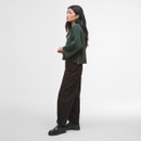 A person with long hair stands sideways against a plain background, wearing a green sweater, black pants, and black shoes, exuding a calm demeanour.