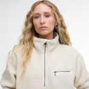 A person with long, wavy blond hair wearing a high-collared, cream-coloured sherpa jacket stands against a plain white background. The expression is neutral.