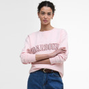 A person in a pink sweatshirt with "Barbour" text stands confidently with crossed arms. Their hair is curly, and they wear gold hoop earrings. The tone is casual.