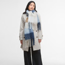 A person wearing a beige trench coat is wrapped in a large, tasselled scarf with blue, white, and tan plaid patterns, conveying a cosy, stylish look.