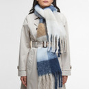 A woman in a beige trench coat, with a blue, white, and brown plaid scarf wrapped around her neck. The scarf's fringes add texture, creating a cosy, stylish look.