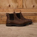Brown leather work boots with black elastic side panels and pull tabs, set against a rustic wooden backdrop. The scene conveys a rugged, durable feel.