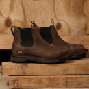 Brown leather work boots with black elastic panels rest on wooden planks. The rugged texture and stitching convey durability and craftsmanship.