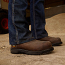 Close-up of a person wearing worn brown work boots and blue jeans, standing on a dusty surface. The boots display a rugged and durable look.