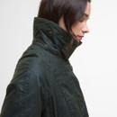 A woman in profile wearing a dark green, high-collared jacket with brass buttons. Her expression is calm, with a minimalist, neutral background.