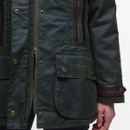 Close-up of a dark green Barbour jacket with gold zippers and button details. The jacket features leather accents and a visible hand at the side.