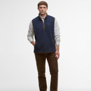 A man stands confidently in a navy vest over a checkered shirt and brown pants against a white background. He has his hand in the vest pocket, conveying a relaxed and casual demeanour.