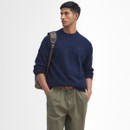 A man in a navy sweater and khaki pants stands confidently against a white background. He has a plaid backpack over one shoulder and his hands in pockets.