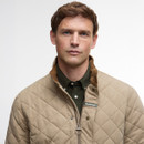 Man in beige quilted jacket with brown collar and green shirt, looking confidently at the camera against a light grey background.