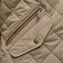 Close-up of a beige quilted fabric with a diagonal zipper pocket and a flap with a round brass snap. The texture appears soft and detailed.