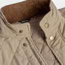 Quilted beige jacket close-up, showing a corduroy-lined collar and brass snap buttons. The texture appears warm and stylish, with a casual, classic feel.