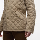 A person wearing a tan quilted jacket with brass buttons and a pocket flap, paired with brown pants, stands against a white background. The mood is casual.