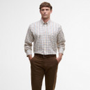 A man wearing a light plaid button-down shirt tucked into brown pants stands confidently with hands in pockets against a plain light background.