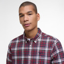 Young person in a red and green plaid button-up shirt looks confidently at the camera against a plain white background, conveying a casual and relaxed tone.